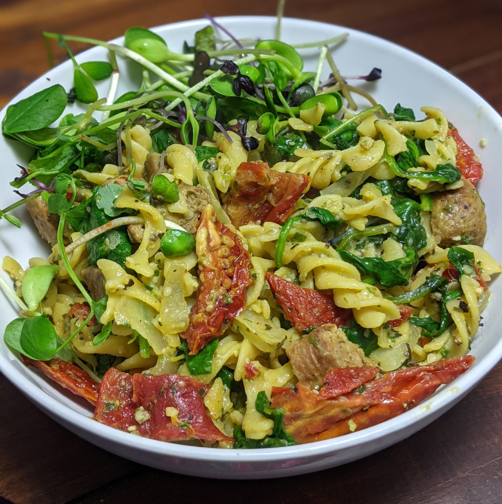 a bowl of sun-dried tomatoes, spinach and pesto pasta with vegan sausage and microgreens